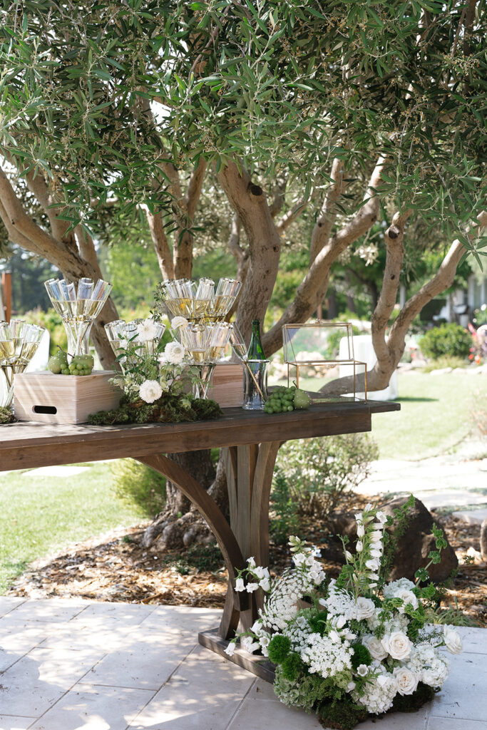 A rustic wooden table outdoors holds floral arrangements, champagne glasses, and decorative greenery, set against a backdrop of trees and grass.