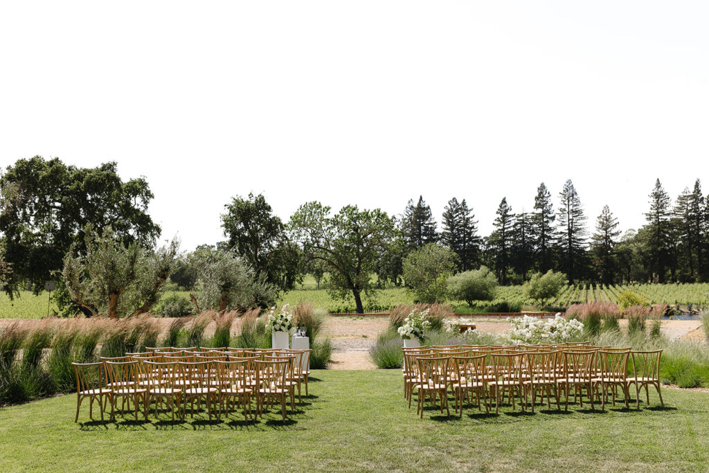 Outdoor wedding setup with wooden chairs arranged on a grassy area, surrounded by tall grasses and trees in the background.