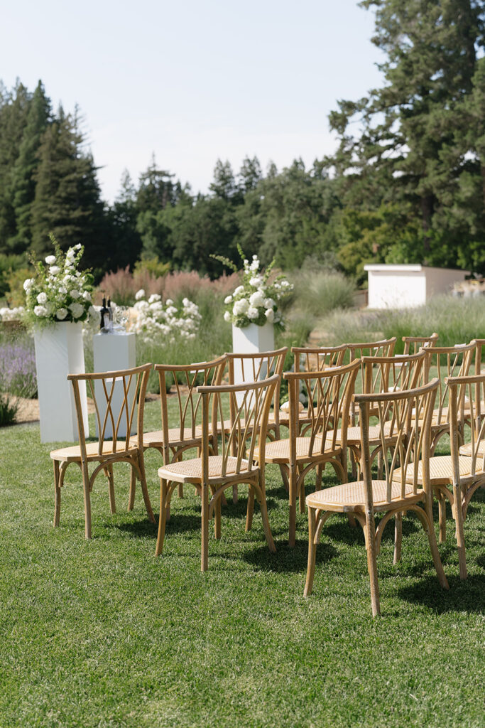 Wooden chairs arranged on grass facing floral arrangements, set outdoors with greenery and trees in the background, suggesting a wedding or event setup.