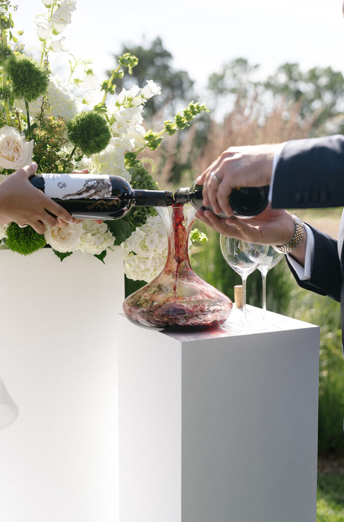 Two people pour red wine from bottles into a glass decanter on a white pedestal, with floral arrangements in the background.