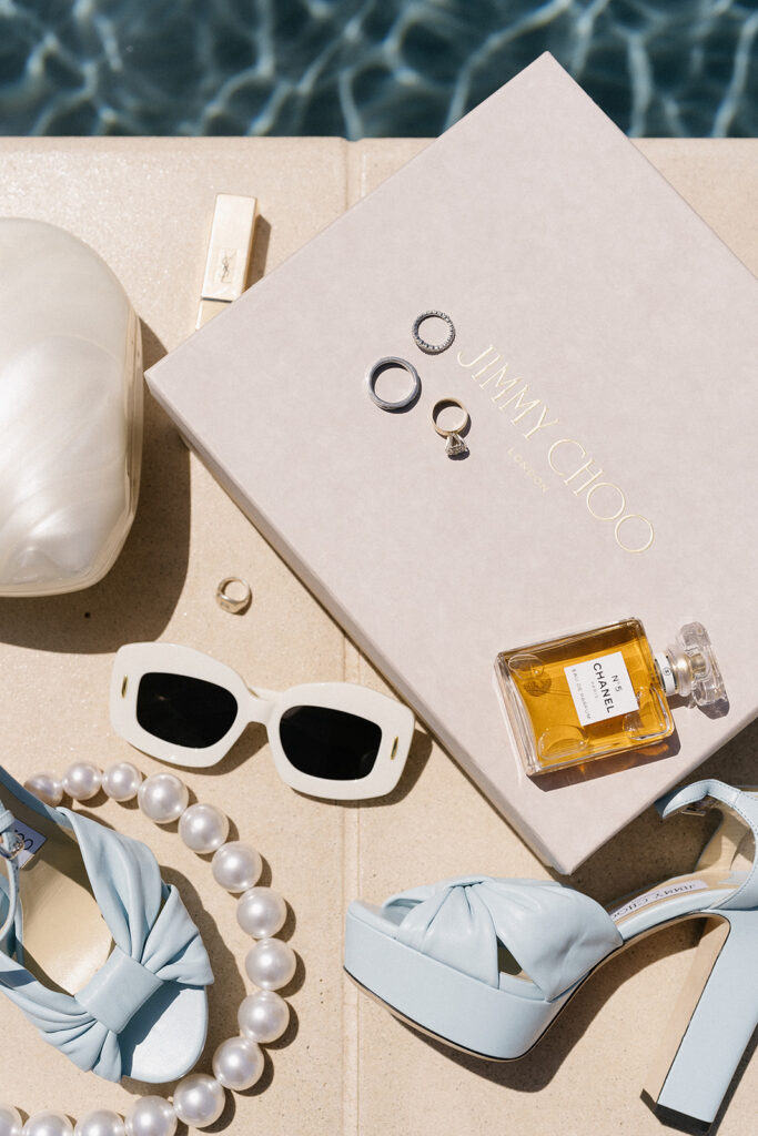 A pair of blue high heels, large pearl necklace, white sunglasses, Jimmy Choo box, Chanel perfume, rings, and a white clutch arranged neatly on a beige surface by a pool.