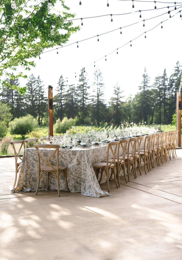 Outdoor dining setup with a long table, floral tablecloth, wooden chairs, and ambient string lights. Surrounded by trees and greenery.