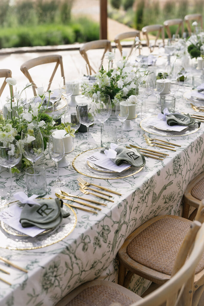 Elegant outdoor table setting with floral tablecloth, white flowers, and greenery centerpieces. Plates with green napkins and monograms; glassware and gold cutlery arranged neatly.