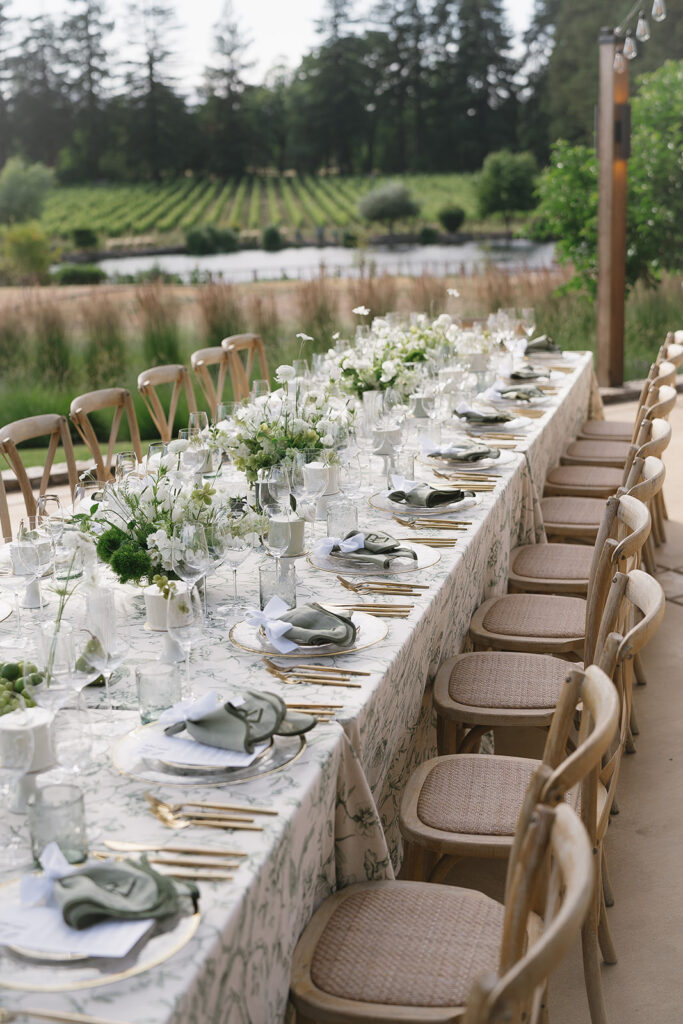A long outdoor dining table is elegantly set with floral centerpieces, glassware, and neatly arranged tableware. Wooden chairs are lined up along both sides, with a view of a vineyard in the background.