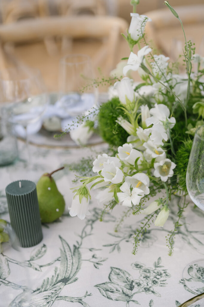 A floral table setting with white bell flowers, a green candle, and a pear on a patterned tablecloth.