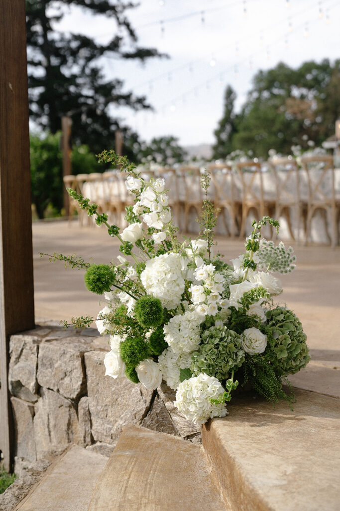 Bouquet of white and green flowers arranged on stone steps, with a blurred outdoor dining area in the background.