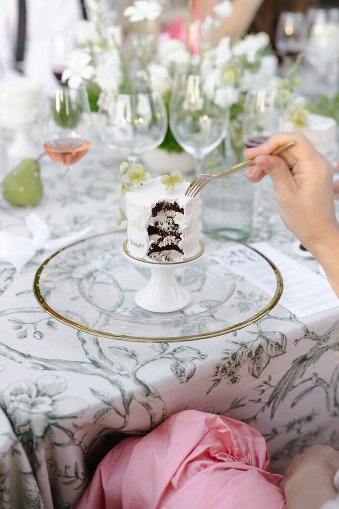 A person holding a fork near a partially eaten white cake on a plate. The table is covered with a floral-patterned cloth, surrounded by wine glasses and a floral arrangement.