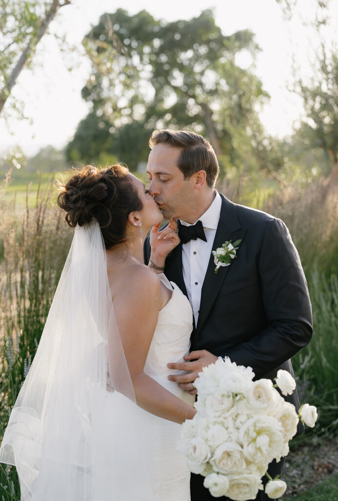 Bride and groom kissing in an outdoor setting, with the groom wearing a black suit and the bride in a white dress, holding a bouquet of white flowers.