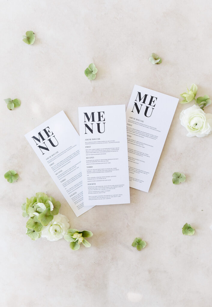 Three white menu cards are arranged on a light surface, surrounded by scattered green leaves and white flowers.
