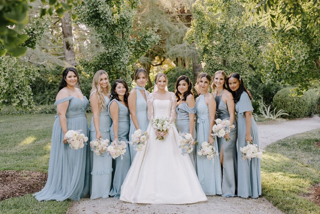 A bride stands with nine bridesmaids. The bride wears a white dress, and the bridesmaids wear matching light blue gowns. They are outdoors, surrounded by greenery.
