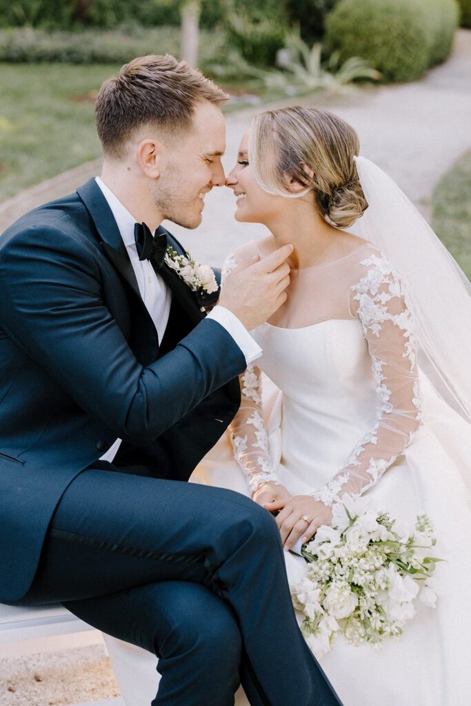 A bride and groom sit closely, smiling at each other. The bride wears a white dress with lace sleeves and a veil, holding a bouquet, while the groom is in a dark suit with a bow tie.
