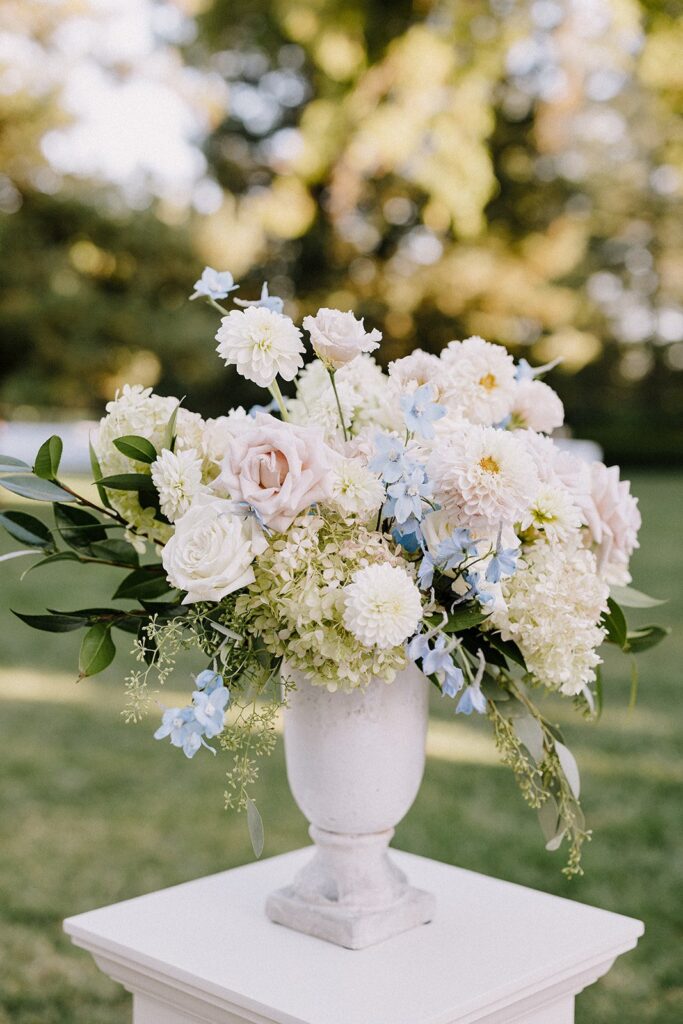 A white vase filled with a bouquet of white and pale pink roses, dahlias, and hydrangeas, accented with light blue flowers, set against a blurred green outdoor background.