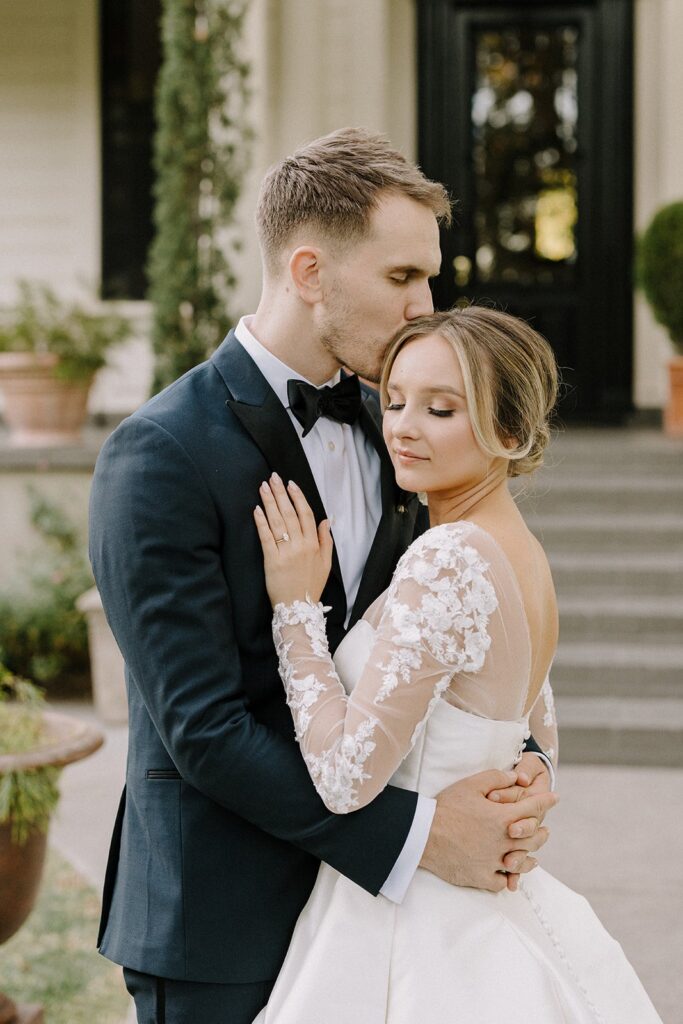 A groom in a black suit kisses the forehead of a bride in a white lace dress as they embrace outdoors on a sunny day.