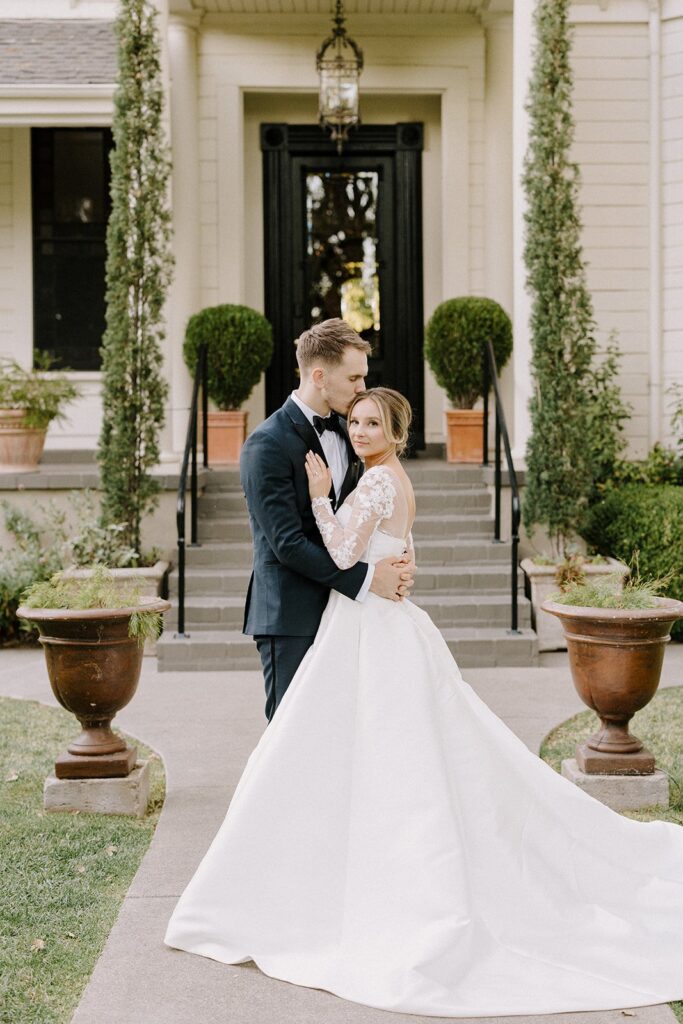 A couple in formal attire embrace in front of a white building with manicured shrubs and steps. The woman wears a white gown, and the man is in a dark suit.