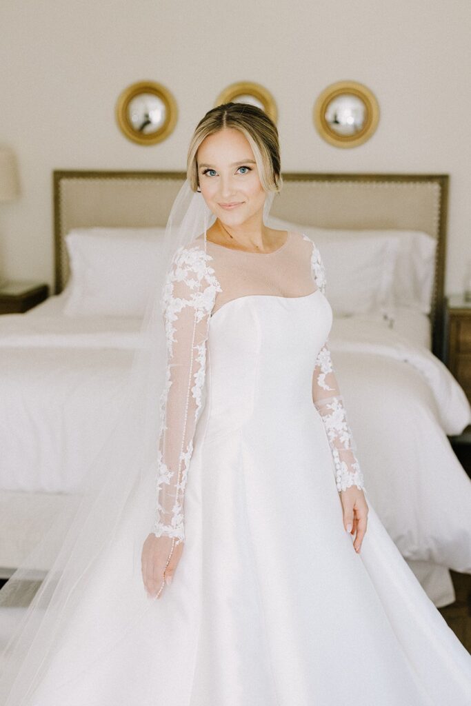 A bride in a white gown with lace sleeves stands in a bedroom, in front of a bed, smiling at the camera. Three circular mirrors are on the wall behind her.