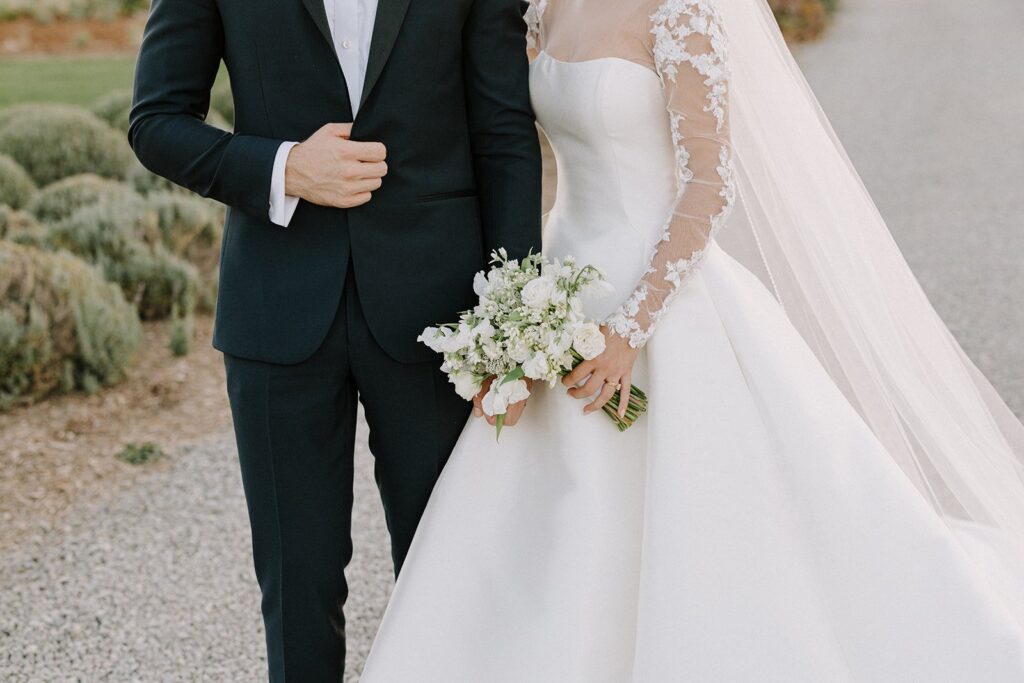 Bride and groom standing together; bride in a white dress holding a bouquet, groom in a black suit.