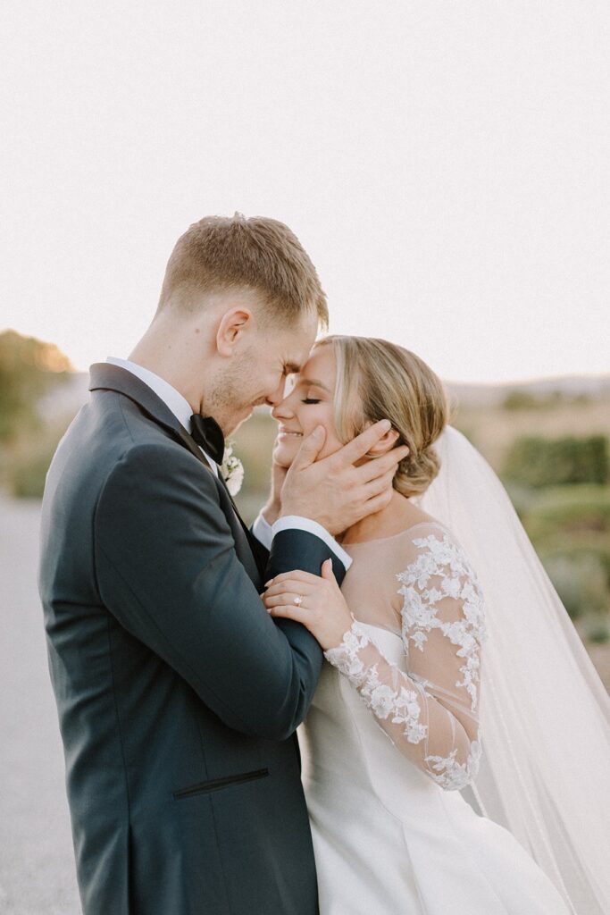 A couple in wedding attire embrace outdoors, smiling gently, with a soft background of greenery and sky.