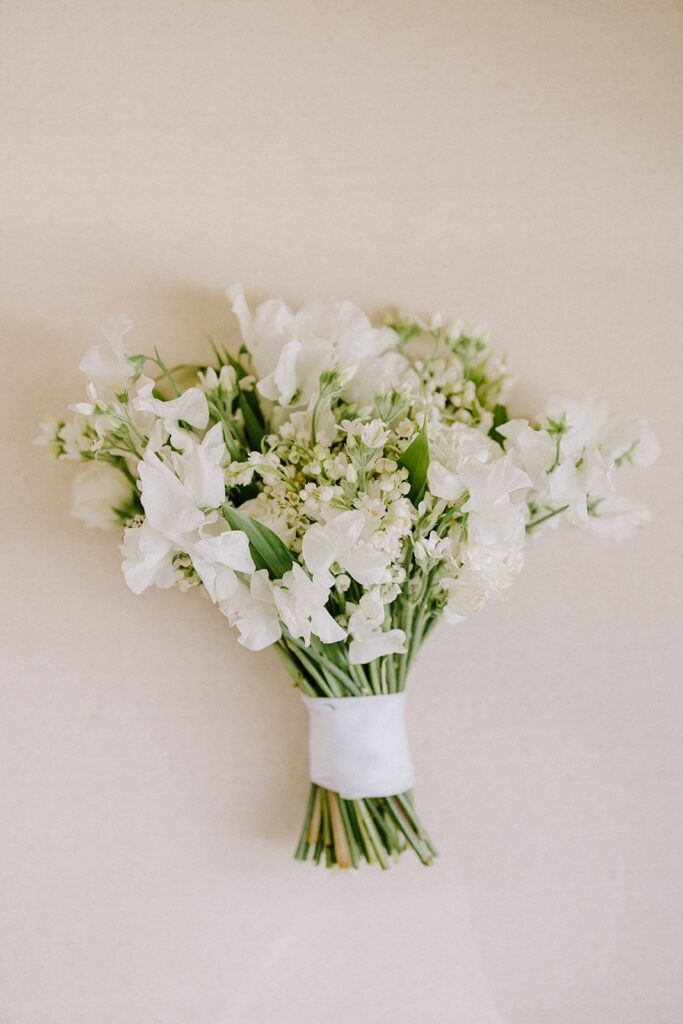 A bouquet of white flowers with green stems and small buds, wrapped with a white ribbon, against a plain beige background.
