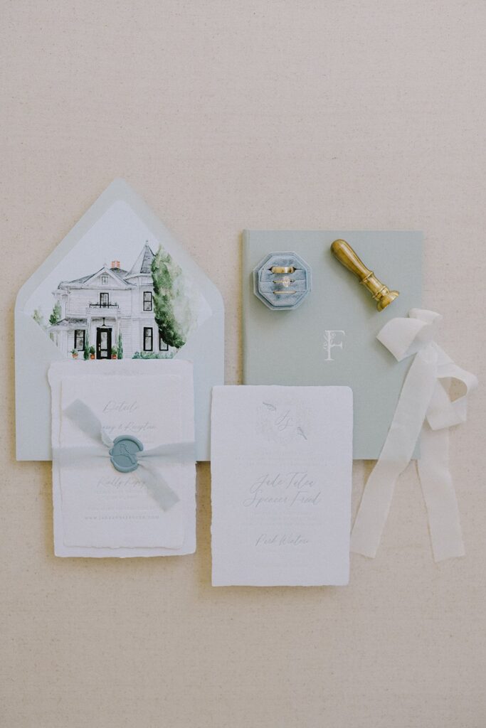 A collection of elegant stationery featuring invitations, an envelope with a house illustration, a wax seal stamp, a ring box, and a ribbon on a beige background.