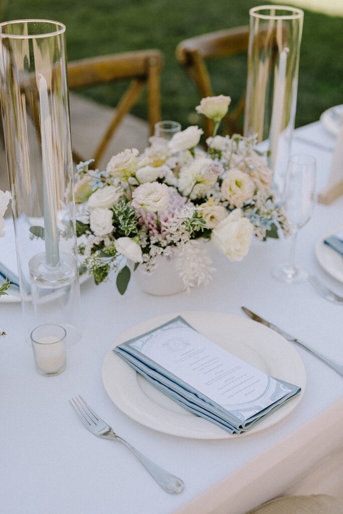 Elegant table setting with white and pastel flowers in a vase, two tall candles, and neatly arranged plates with folded menus on a white tablecloth.