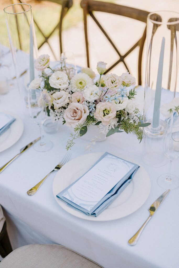 Elegant table setting with plates, blue napkins, menu cards, white flowers, and blue candles on a white tablecloth.