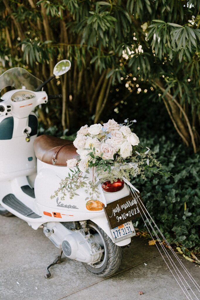 White scooter decorated with a floral arrangement on the seat, a small "Just Married 9-10-2023" sign at the back, parked near green foliage.