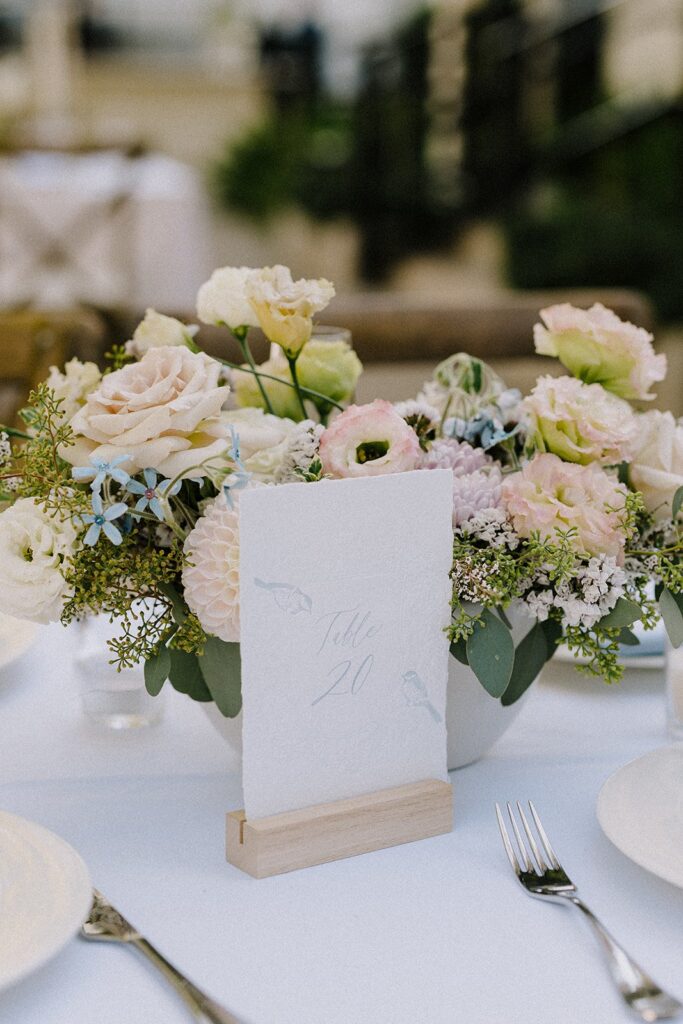 A floral centerpiece with pastel flowers and greenery on a white tablecloth. A card with "Table 20" is placed in a wooden holder. Plates and a fork are visible.