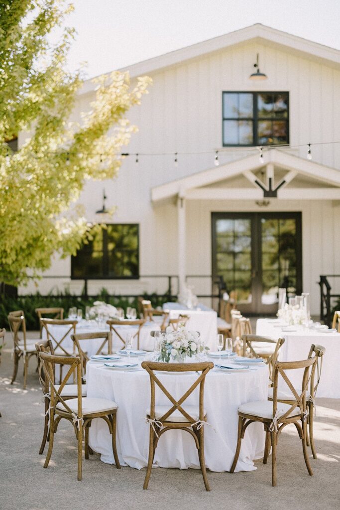 Outdoor wedding reception setup with round tables covered in white tablecloths, wooden chairs, and floral centerpieces in front of a white building.