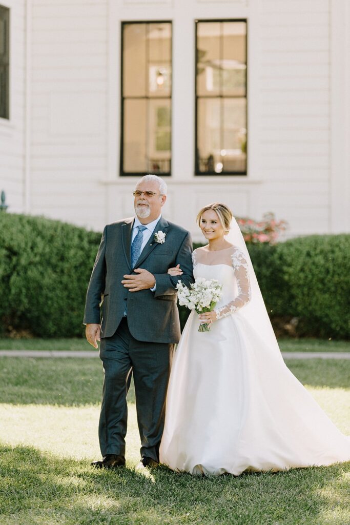 A bride in a white gown and veil walks arm-in-arm with an older man in a suit, holding a bouquet of flowers. They are outside, with greenery and a white building in the background.