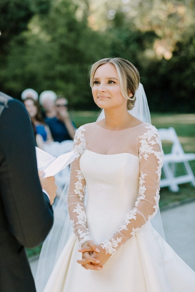 A bride in a white dress with lace sleeves stands outdoors, hands clasped, facing a person holding a paper. White chairs and greenery are in the background.