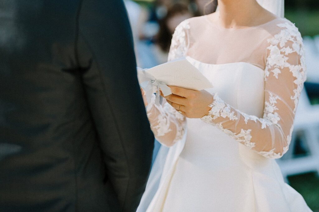 Bride in a white dress reads vows from paper while standing next to a groom in a black suit at an outdoor ceremony.