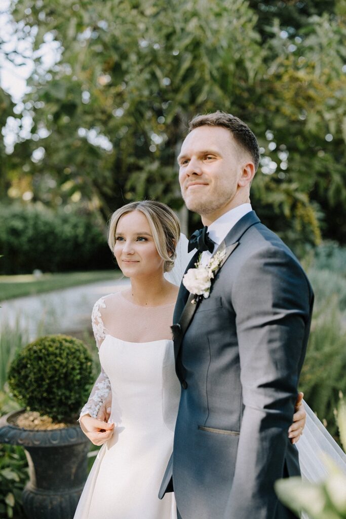 A bride and groom stand outdoors, embracing and looking ahead. They are dressed in formal wedding attire, surrounded by greenery.