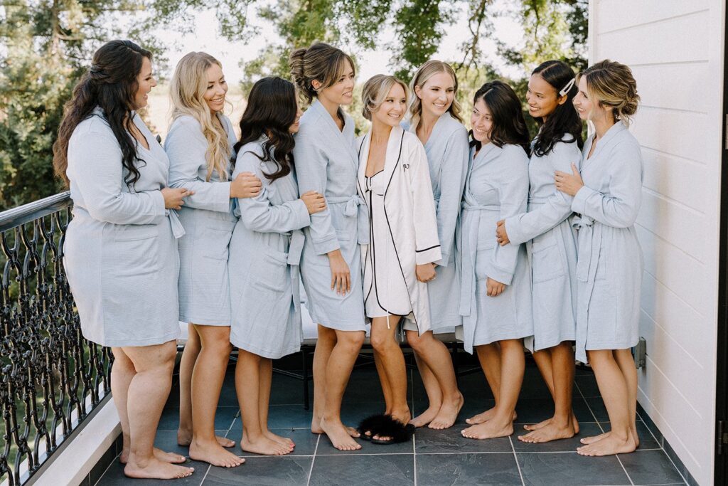 A group of women wearing matching bathrobes stand together on a balcony, smiling. Trees and a railing are visible in the background.