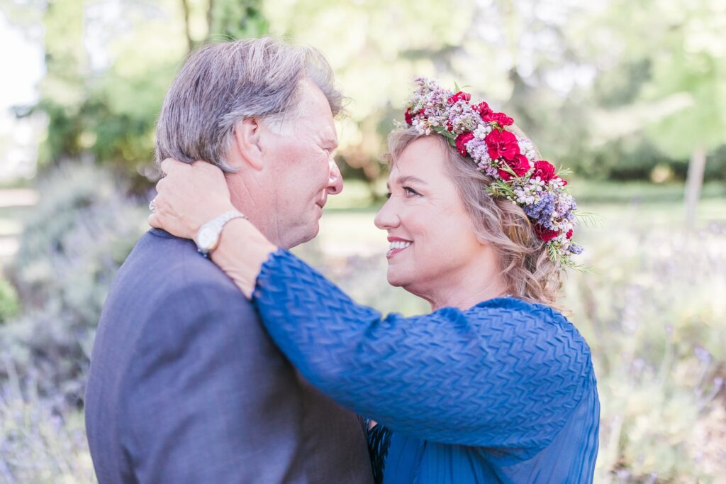 A couple stands outdoors, facing each other closely. The woman with a flower crown smiles at the man, who wears a gray jacket. The background is blurred greenery.