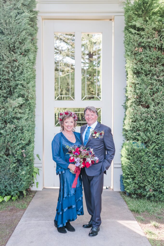 A couple stands in front of a white door flanked by tall greenery. The woman holds a bouquet, and both wear formal attire with floral accents.