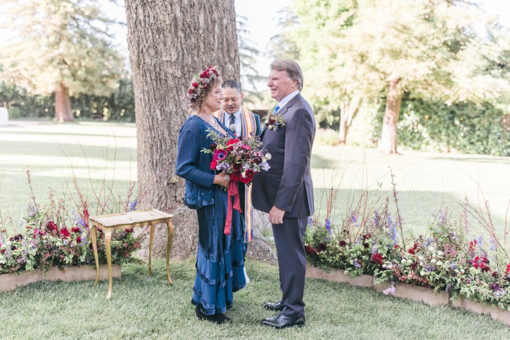 A couple stands together outdoors during a wedding ceremony, with a person officiating. They are surrounded by flowers and greenery.