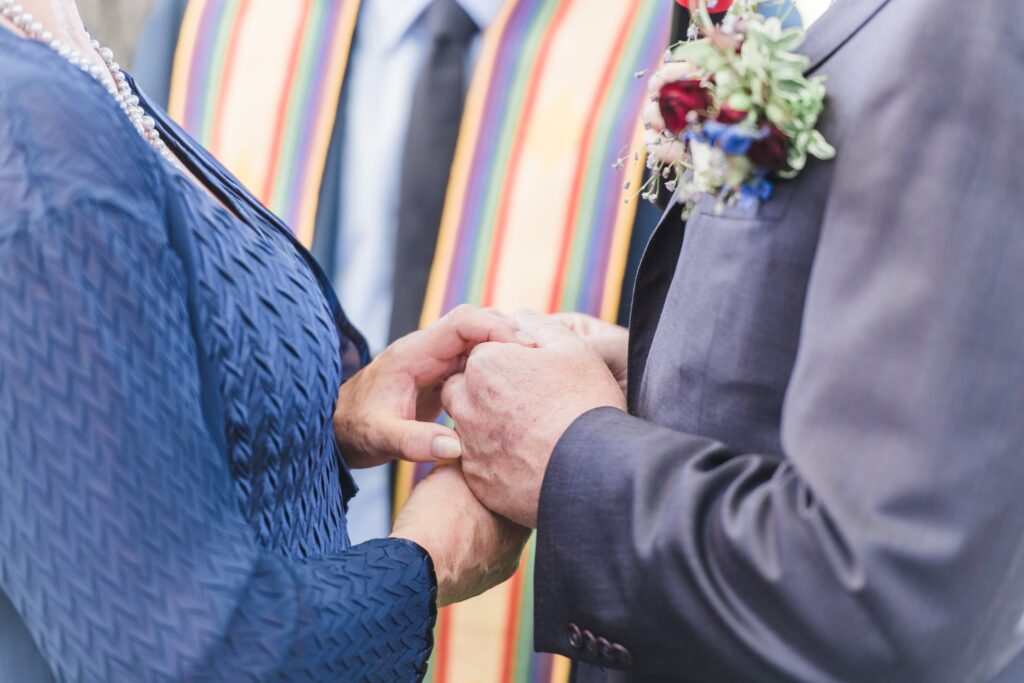 Two people holding hands during a ceremony, with one wearing a dark suit and floral corsage, and the other in a blue textured garment. A person with a colorful stole stands nearby.