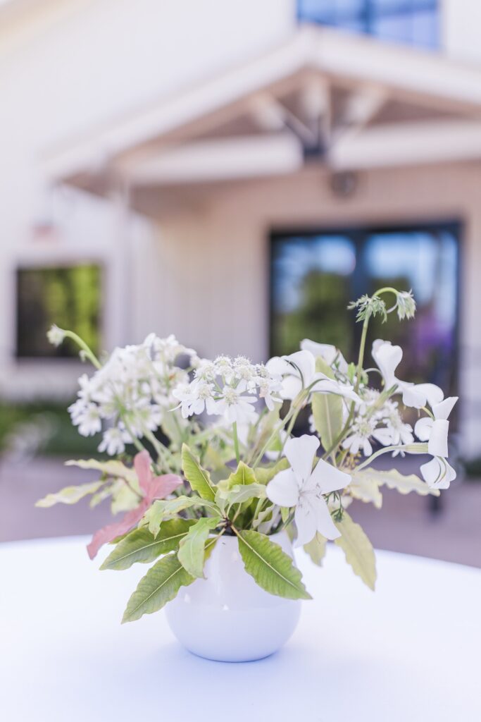 A white vase with white flowers and green leaves on a table, set outdoors near a modern house.