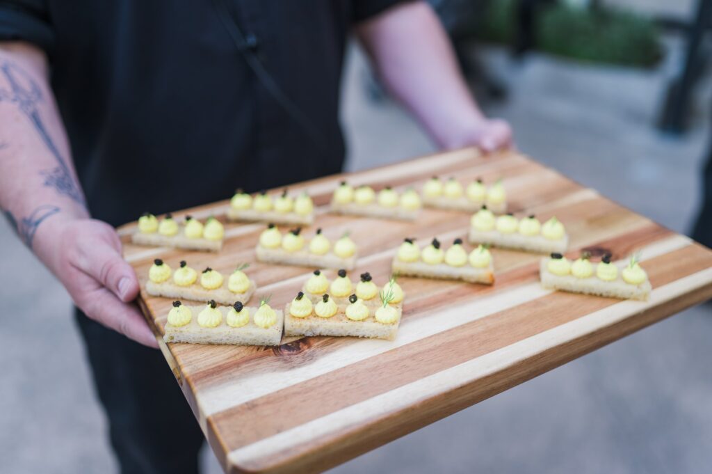 Person holding a wooden tray with multiple rectangular appetizers topped with yellow cream and small garnishes.