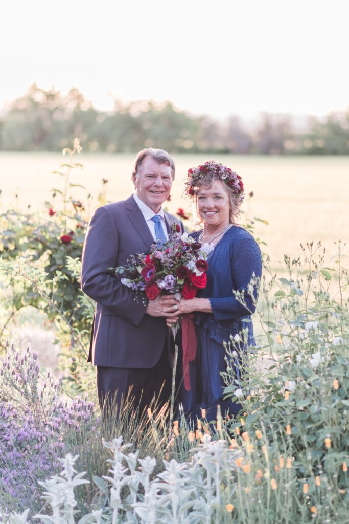 A couple stands outdoors, smiling and holding a bouquet. The man is in a suit, and the woman wears a blue dress with a floral crown. They are surrounded by greenery and flowers.