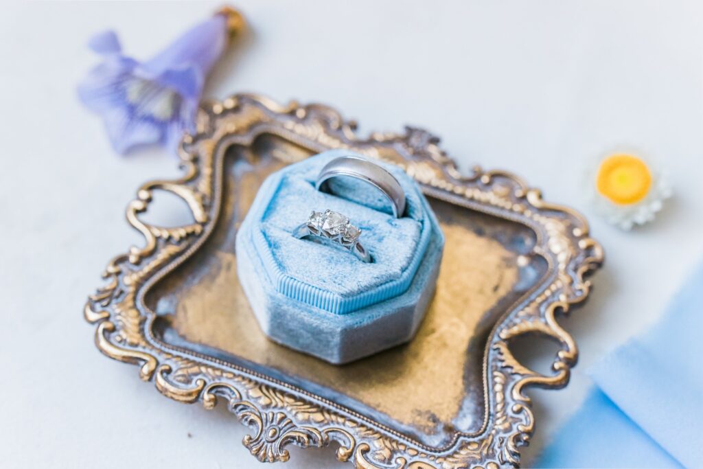 A diamond ring in a light blue velvet box on an ornate metal tray with a blue flower and a yellow-centered white flower nearby.