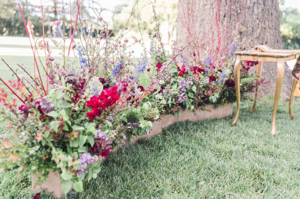 A floral arrangement with red, purple, and green flowers is placed beside a tree. A small gold table stands on green grass nearby.