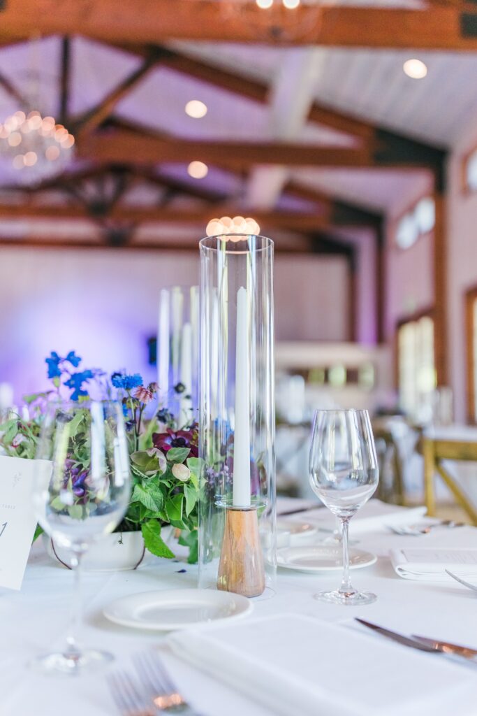 Elegant table setting with a tall glass-enclosed candle, wine glasses, and purple flower centerpiece, under wooden beams in a softly lit room.