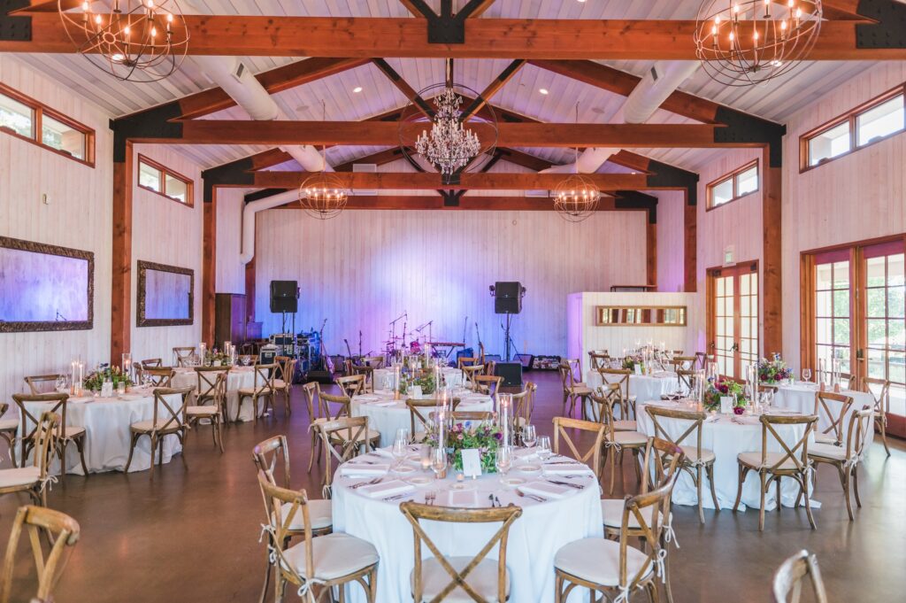 Spacious event hall with round tables set for dining, floral centerpieces, a wooden ceiling with beams, chandeliers, and a stage with musical instruments.