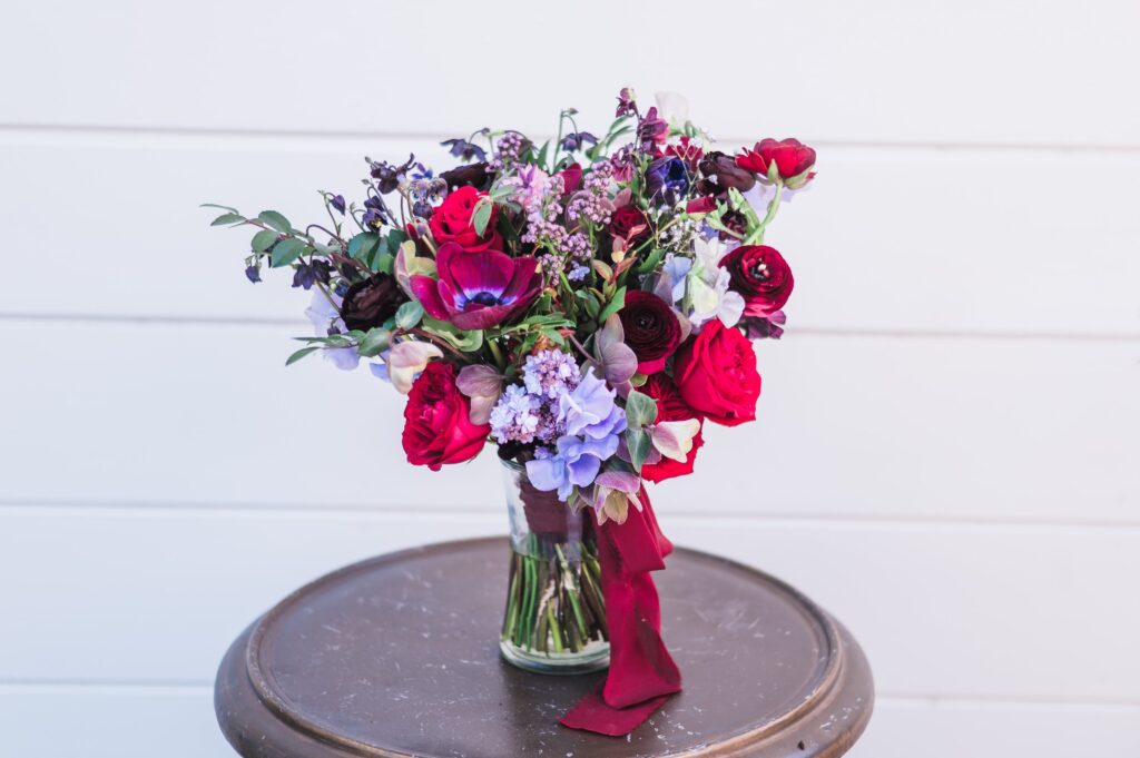 A bouquet of red, pink, and purple flowers with greenery in a glass jar, placed on a round brown table against a white background.