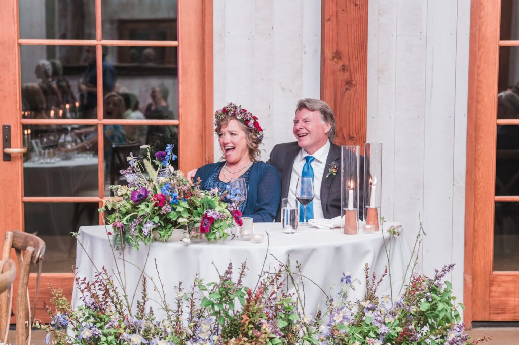 A couple sits at a decorated table, smiling and laughing. The table is adorned with flowers, and candles are lit. They are indoors, near large windows.