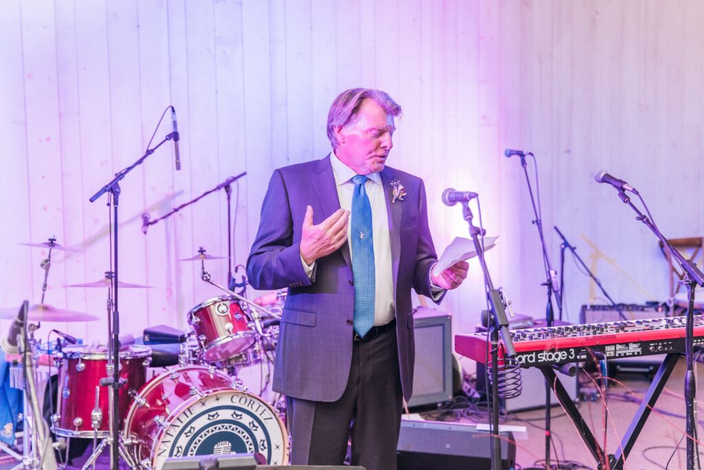 A man in a suit speaks into a microphone, holding a paper. Musical instruments, including a drum set and keyboard, are in the background.