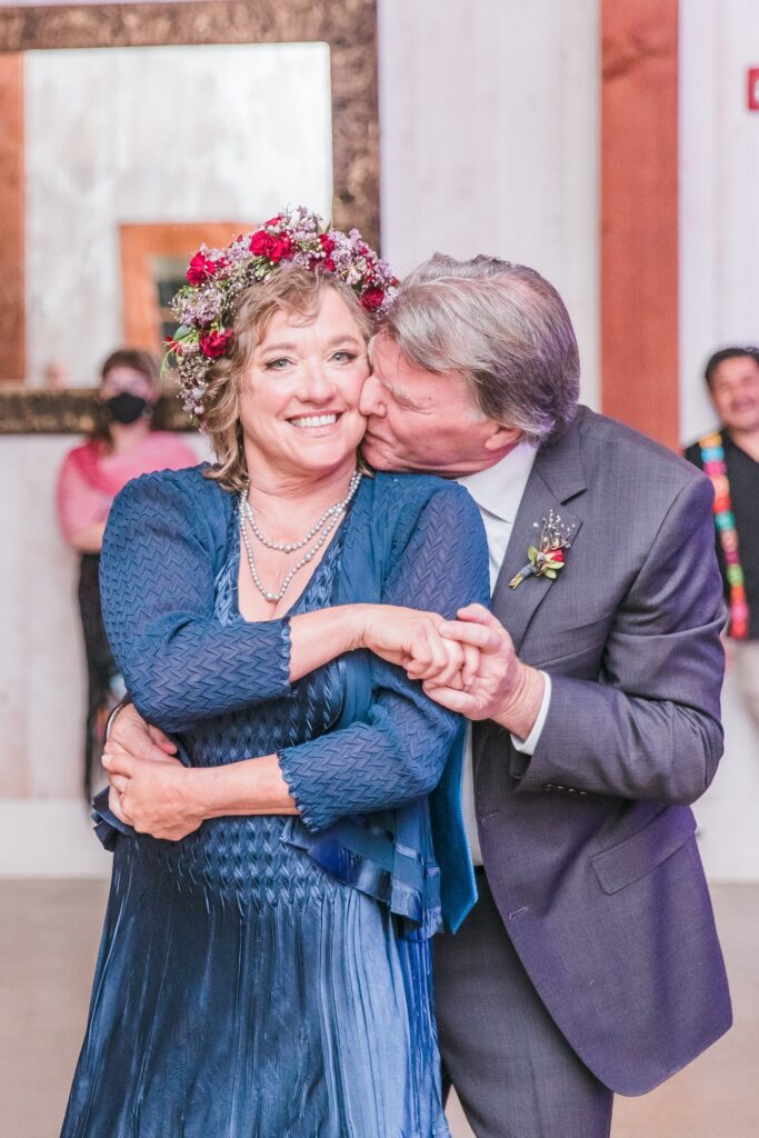An older couple embraces and smiles; the man kisses the woman's cheek. She wears a floral crown and blue dress, and he is in a suit with a boutonniere. People and a mirror are in the background.