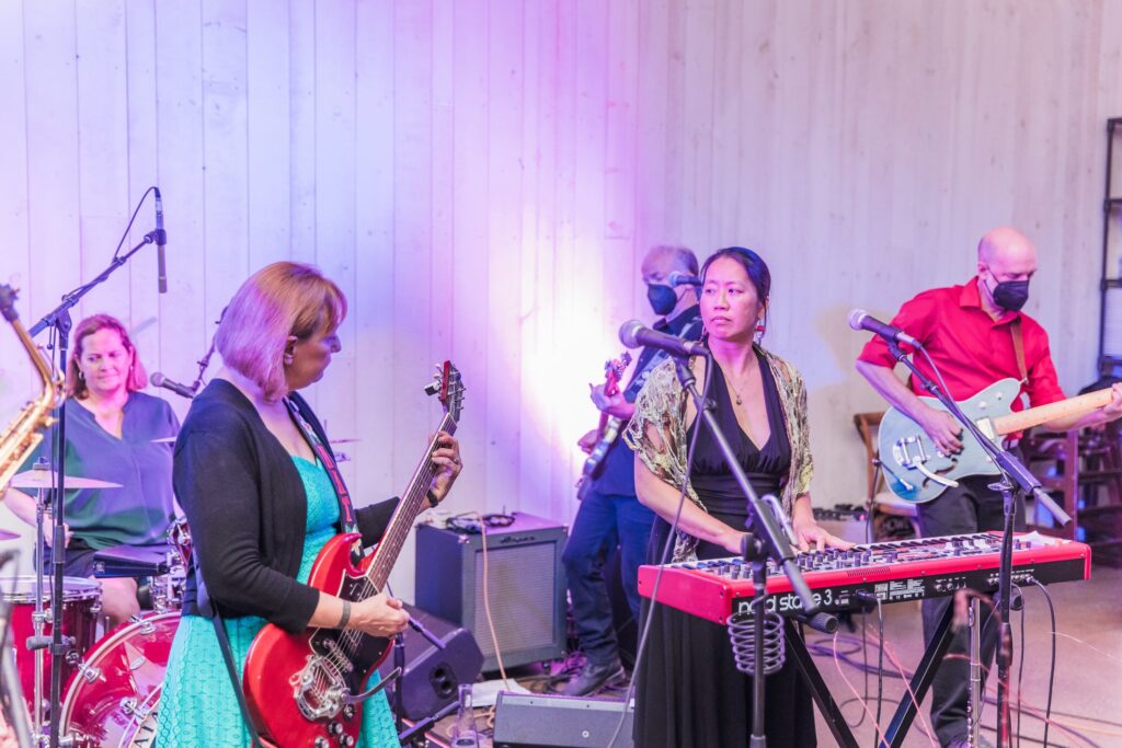 A band performing on stage with guitars, keyboard, and drums. Musicians are wearing casual clothing, and one is wearing a mask. The stage is lit with pink and purple lights.