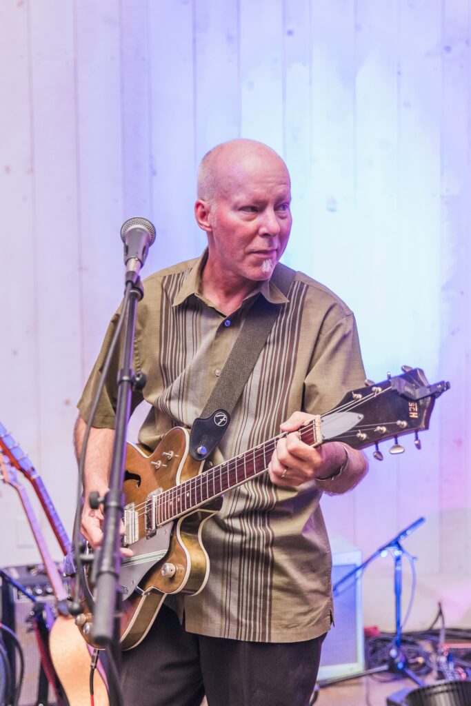 Man playing an electric guitar on stage with a microphone in front, wearing a brown shirt.